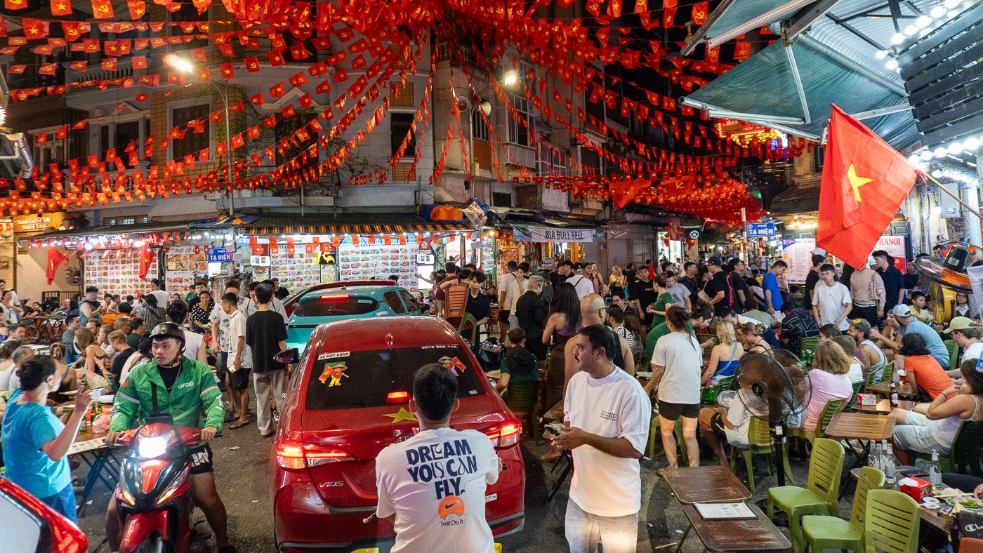 Busy street scene in Hanoi with traffic, people, and street food stalls creating a dense, everyday moment