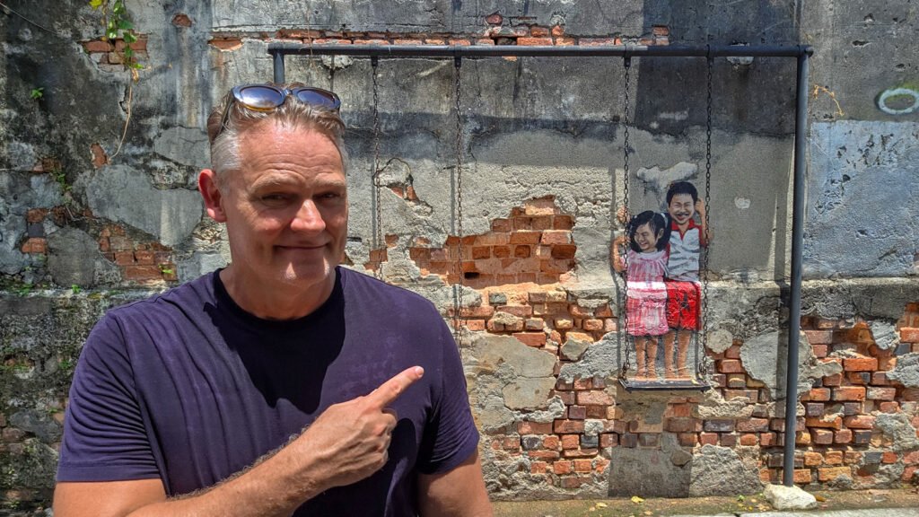 Martin pointing at the famous street art mural of the children on a swing in George Town, Penang