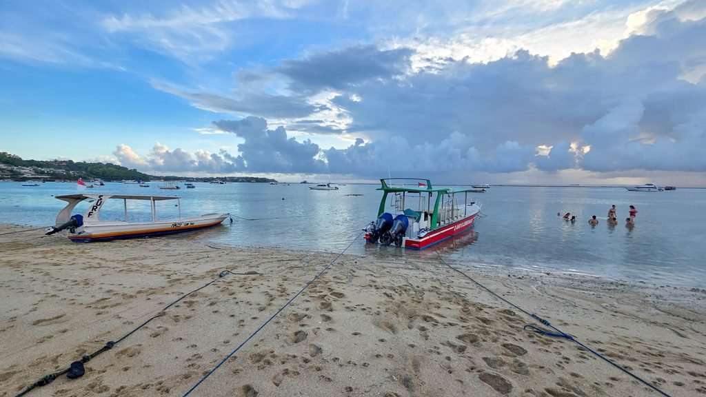 Two boats on the beach at sunset in Nusa Lembongan, with people swimming in the background.