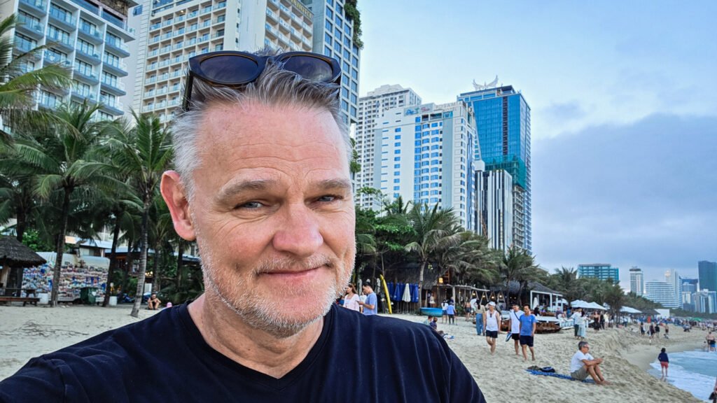 Martin at My Khe Beach in Da Nang, Vietnam, with palm trees, beachfront hotels, and the ocean in the background.