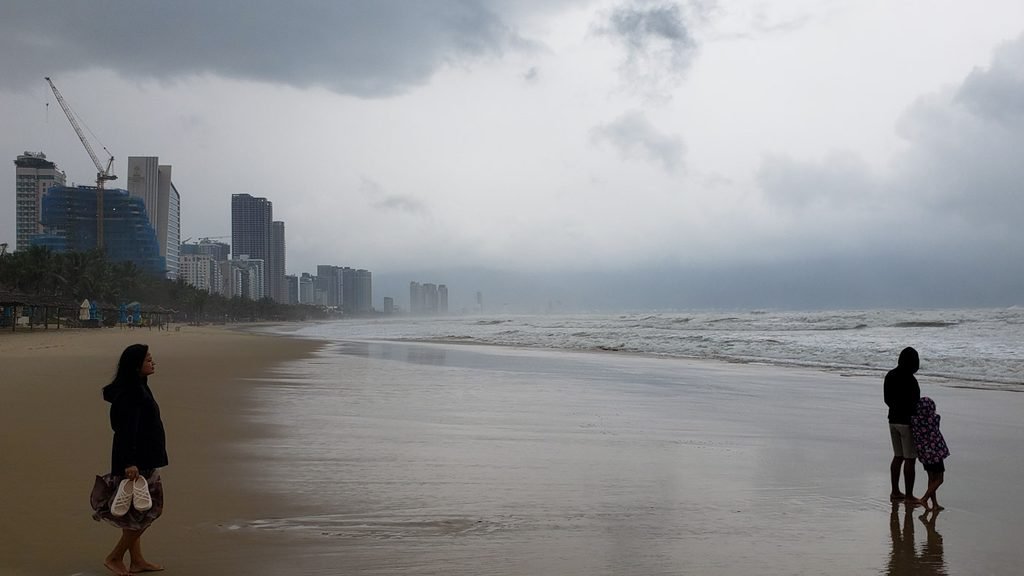 People standing on My Khe Beach in Da Nang, Vietnam, beneath grey storm clouds, with rough waves and beachfront buildings in the distance.